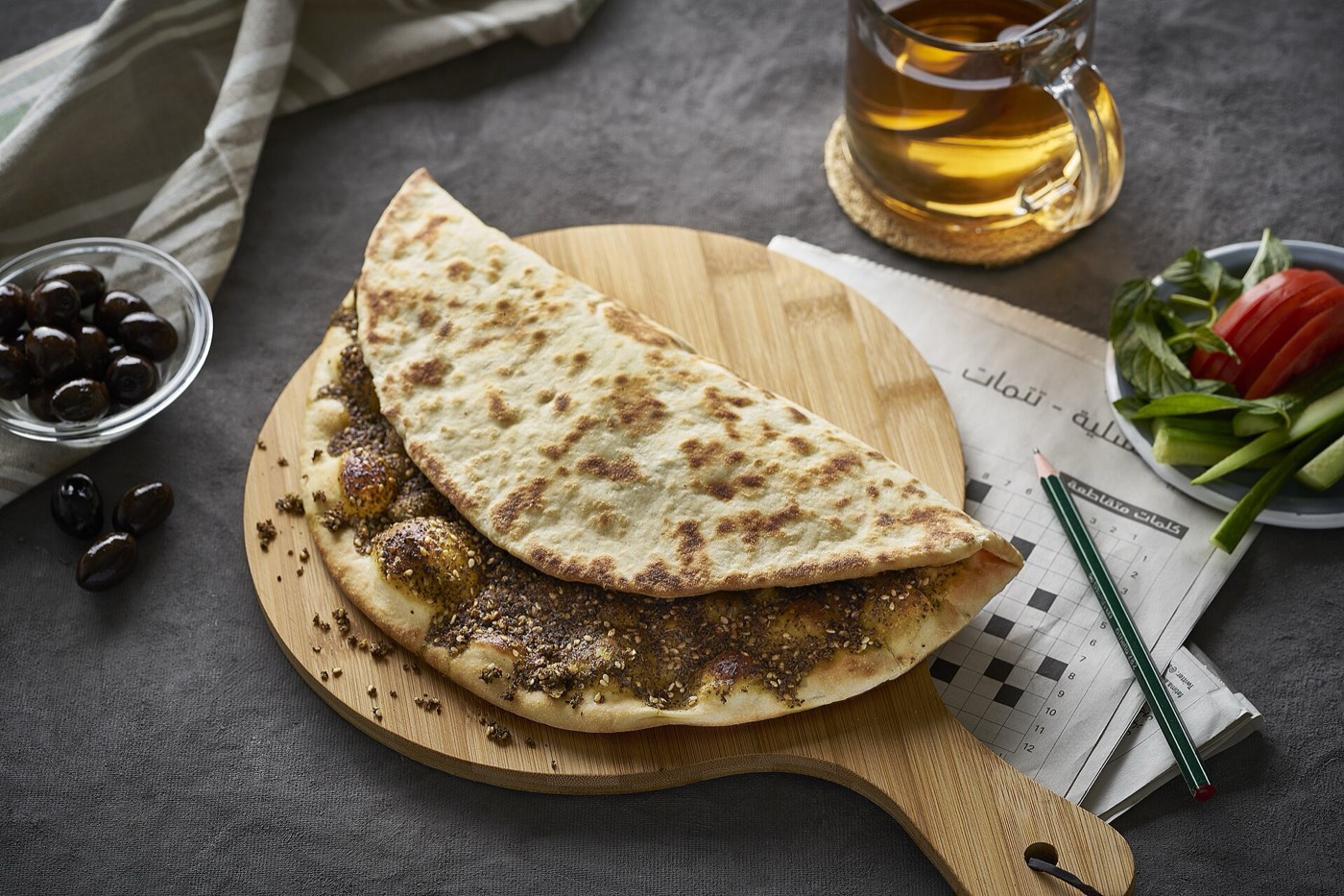 A zaatar manakish folded in half on a wooden board, with tea, olives and fresh vegetables